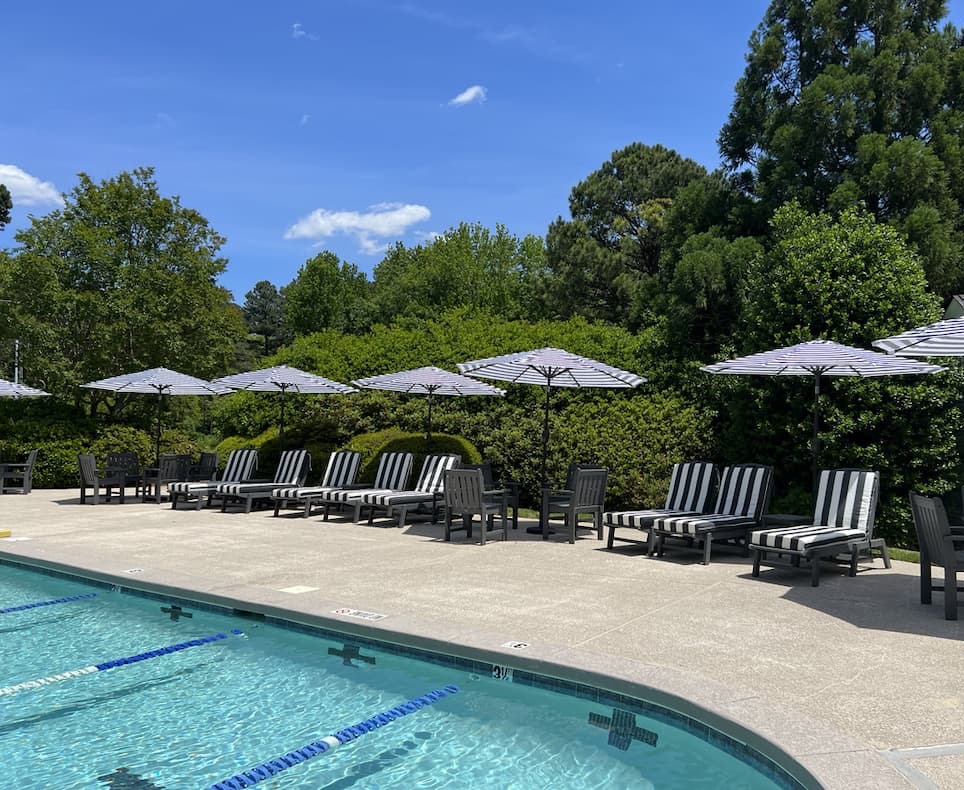 Outdoor swimming pool surrounded by black-and-white striped lounge chairs and matching umbrellas. The sunlit pool deck is bordered by lush green trees and shrubs beneath a bright blue sky with a few scattered clouds, creating a peaceful, resort-like setting.