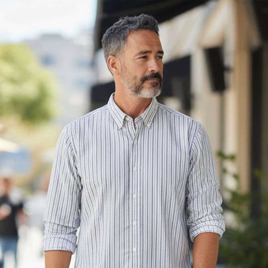 Middle-aged man with short salt-and-pepper hair and a trimmed beard wearing a light striped button-down shirt, walking outdoors and looking to the side.