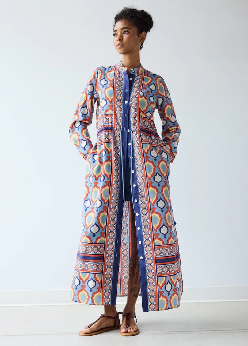 Person standing against a plain wall wearing a long, colorful patterned button-front dress or duster with brown sandals, looking off to the side.