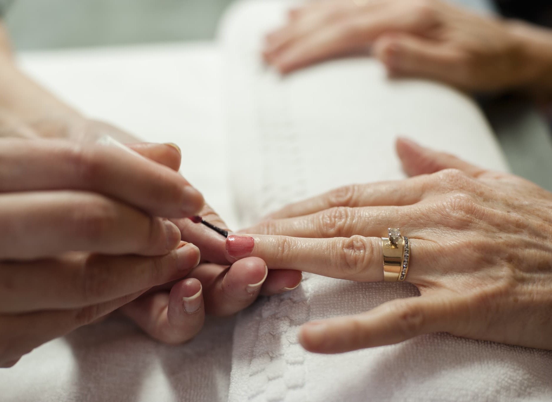 A nail technician carefully applies pink polish during a manicure treatment. The close-up view shows hands resting on a soft towel as one nail is painted with precision, while a simple gold ring adds an elegant touch. The softly blurred background creates a calm spa atmosphere focused on self-care, grooming, and relaxation.