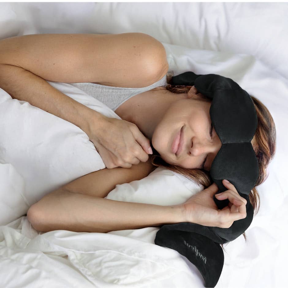 Woman resting in bed wearing a soft black weighted eye mask. Fearrington Village.