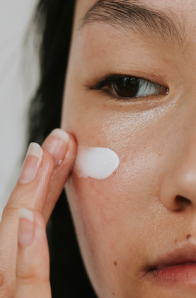 Close-up of a person applying a white facial cream to their cheek with their fingertips. The image focuses on smooth, glowing skin and the eye area, highlighting the texture of the moisturizer as it is gently blended into the face. The neutral background and tight crop create a clean, minimalist skincare aesthetic.