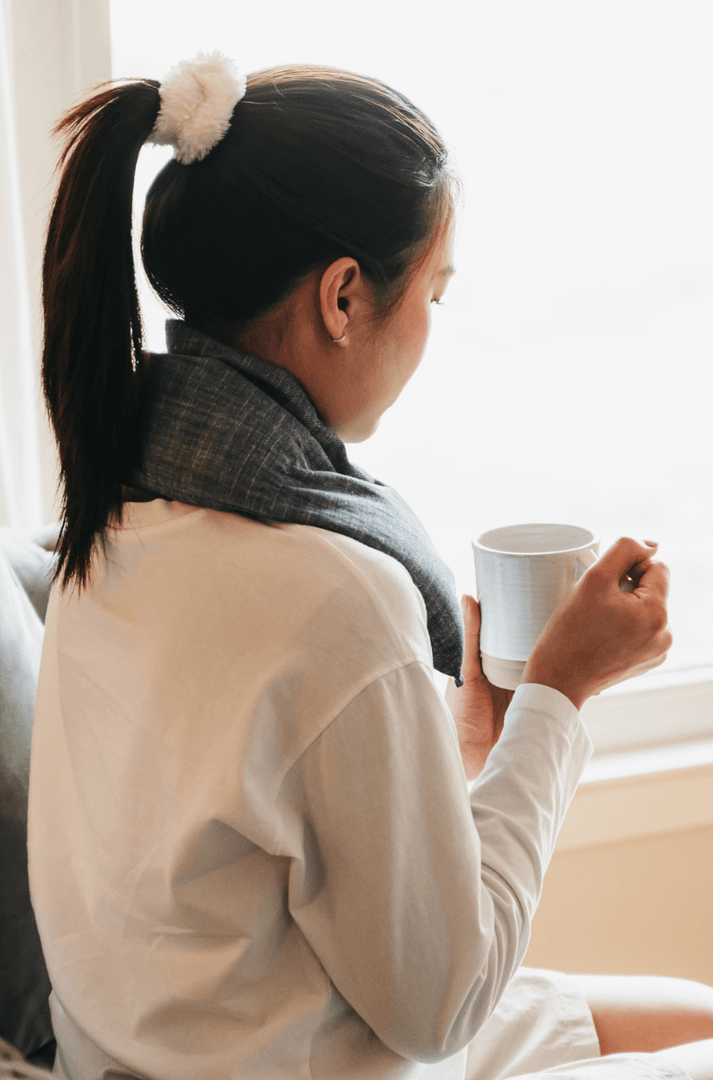 woman viewed from behind holding a mug of tea, her neck draped with a neck wrap