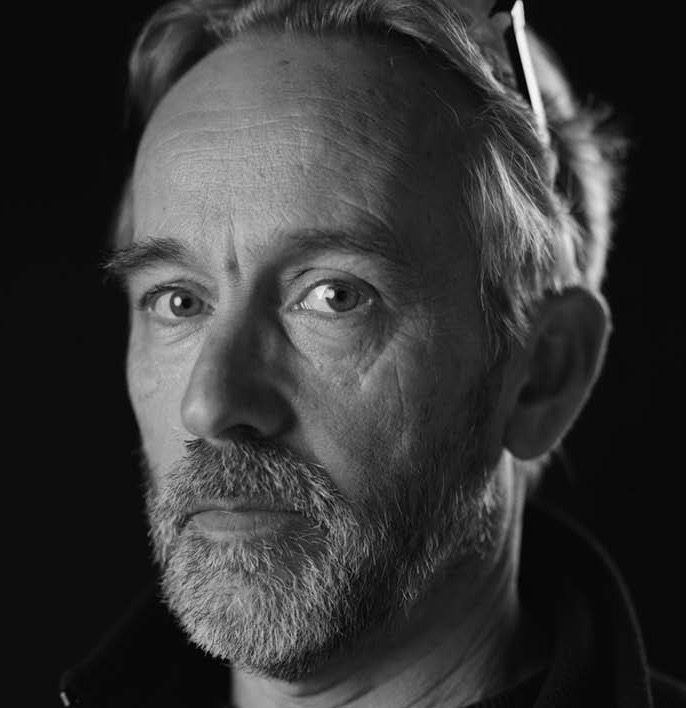 Black-and-white close-up portrait of a bearded man looking toward the camera against a dark background. Fearrington Village.