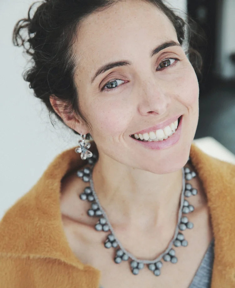 Close-up portrait of a smiling woman with natural makeup and bright eyes, looking directly at the camera. She wears a beaded statement necklace and matching earrings, with a warm-toned coat draped around her shoulders against a softly blurred indoor background.