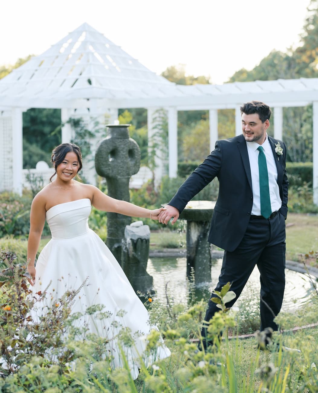 Bride in a strapless white gown and groom in a dark suit hold hands while walking through a garden beside a fountain, with a white pergola in the background.