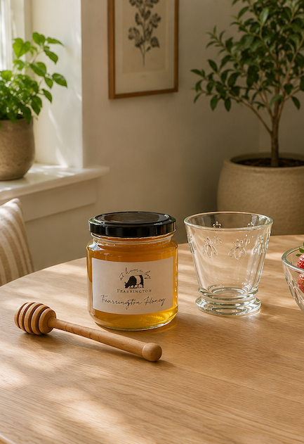 Jar of Fearrington Honey displayed with a wooden honey dipper and bee-etched glass on a sunlit breakfast table, with fresh strawberries and potted plants in a warm Scandinavian-inspired kitchen. Fearrington Village.