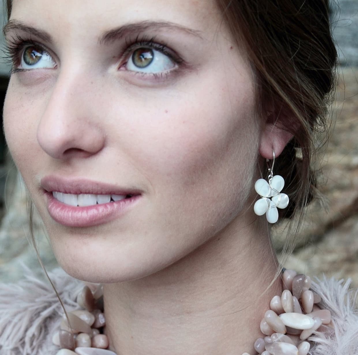 Close-up portrait of a woman with green eyes and soft, natural makeup, looking slightly upward. She wears white floral drop earrings and a chunky, pale stone necklace, with a textured, neutral-toned background.
