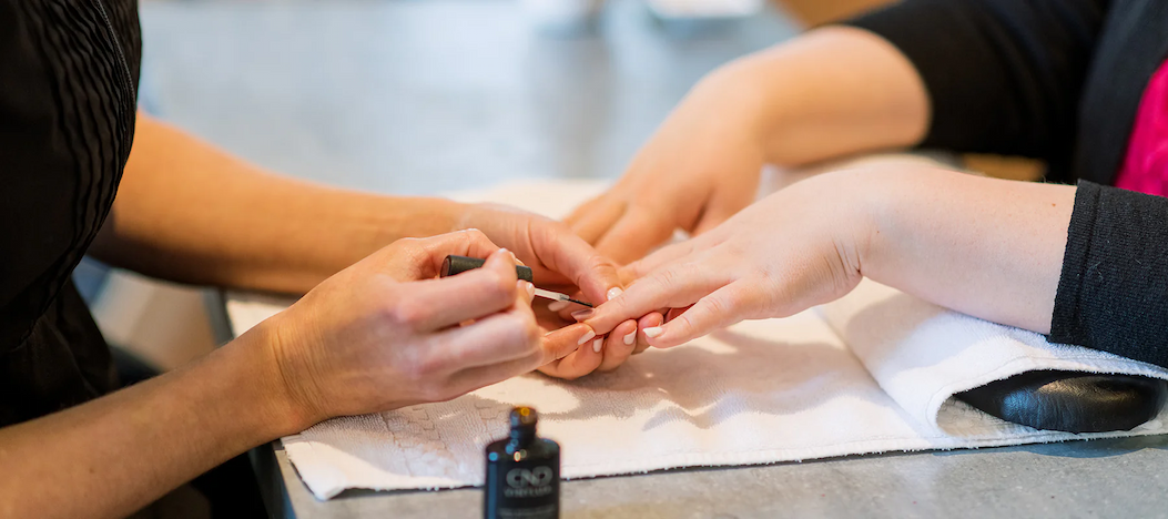 A manicure in progress as a technician carefully paints a client’s fingernails while the client’s hands rest on a folded white towel at a salon table.