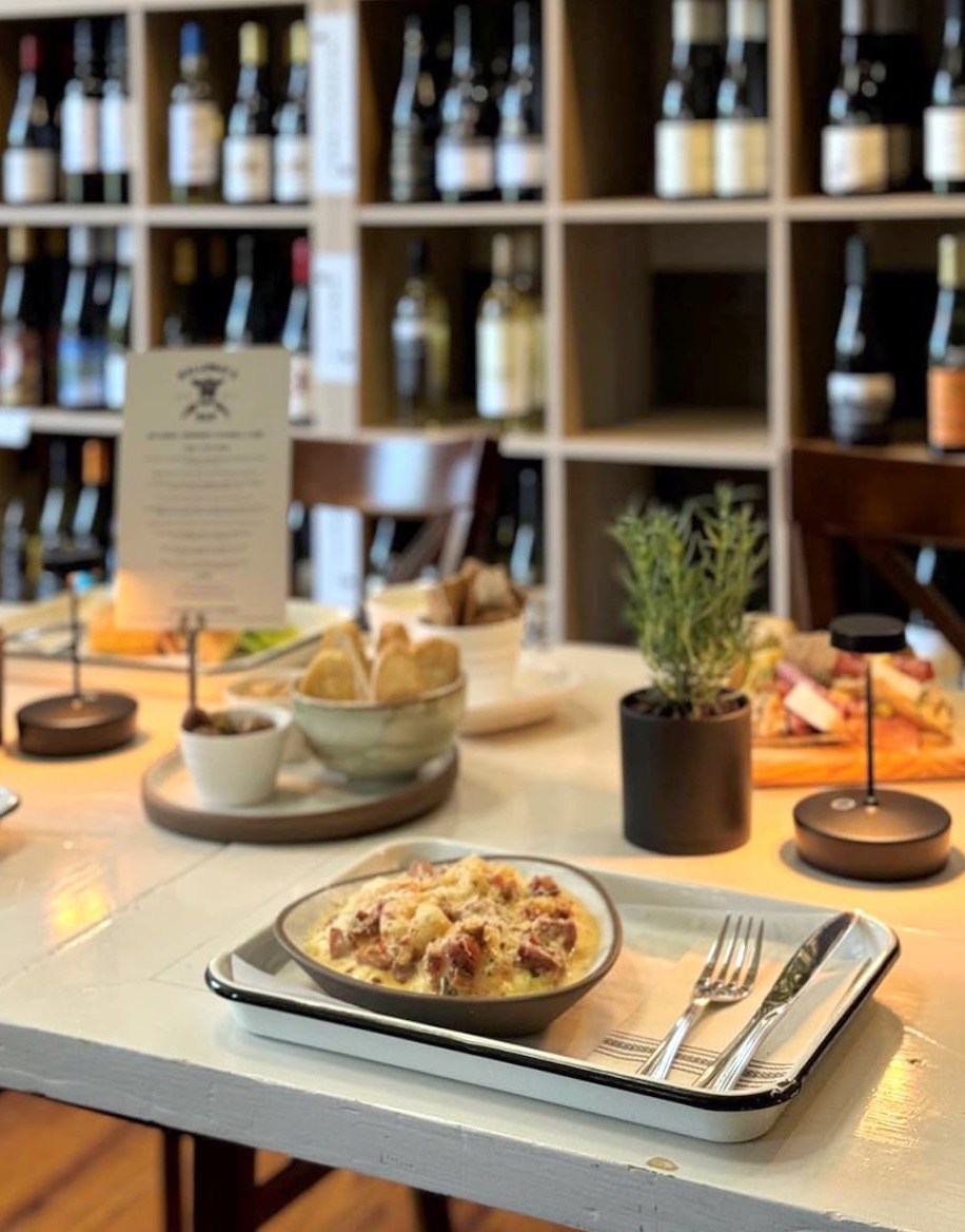 A table set with plated food, cutlery, small potted herbs, and wine glasses sits in front of shelves filled with various bottles of wine in a cozy restaurant setting. Fearrington Village