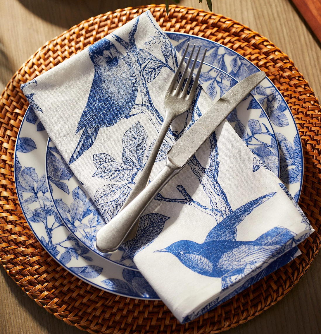 Place setting with a woven rattan charger, blue-and-white botanical dinner plate, folded napkin with bird and leaf pattern, and silver fork and knife on top.