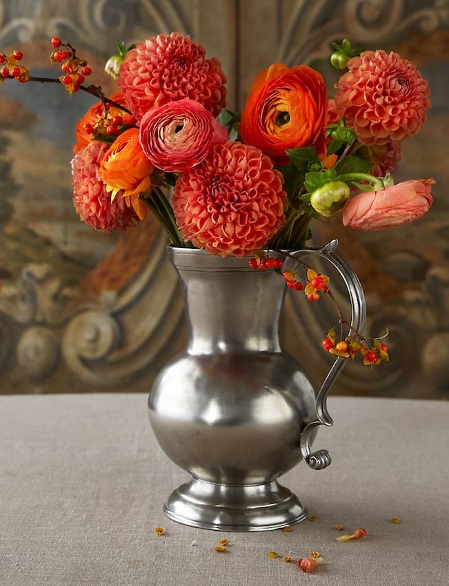 A silver pitcher filled with orange and coral flowers sits on a gray tablecloth, with a few petals scattered beside it. Ornate, painted wooden panels serve as the background. Fearrington Village