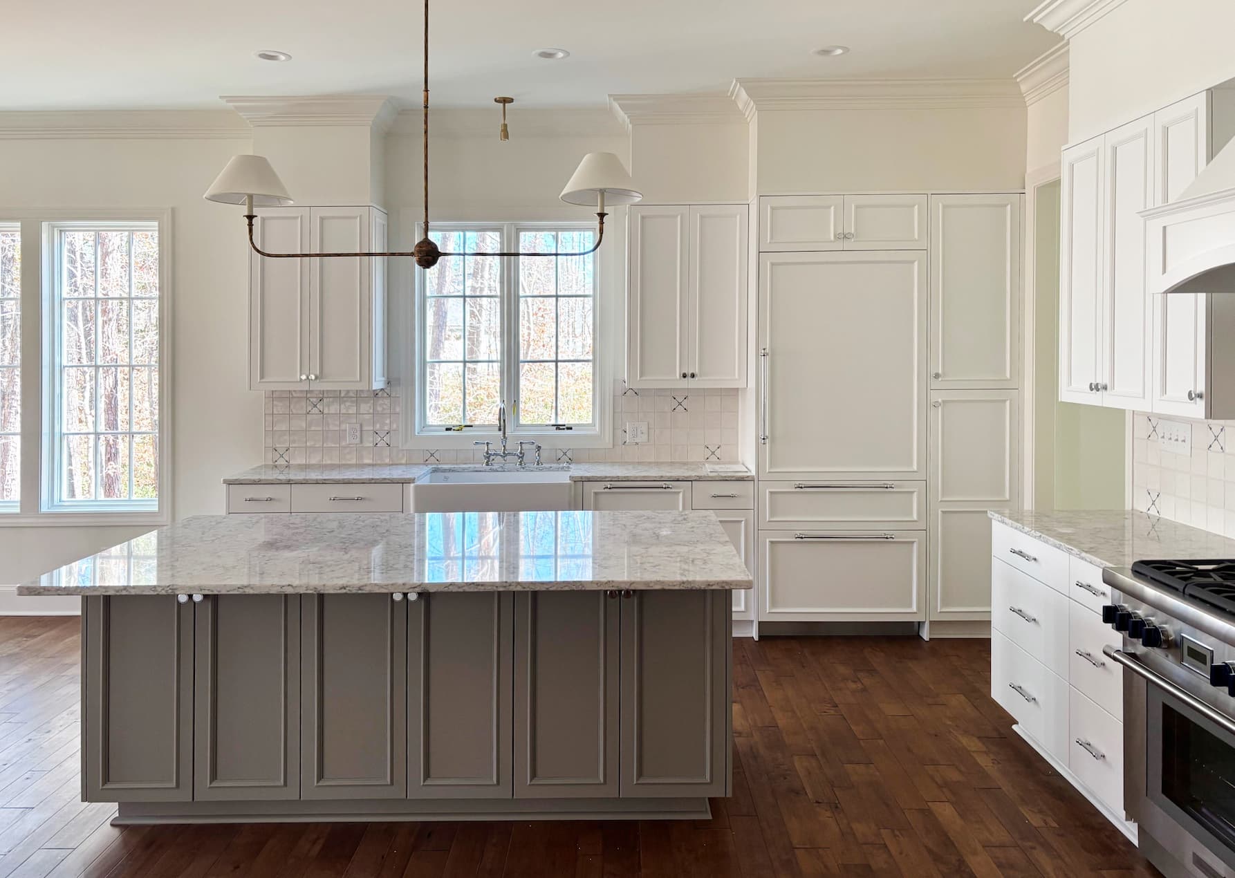 Bright, spacious kitchen with white shaker cabinets, a large gray island topped with marble, farmhouse sink beneath tall windows, warm hardwood floors, and a modern two-light pendant overhead.