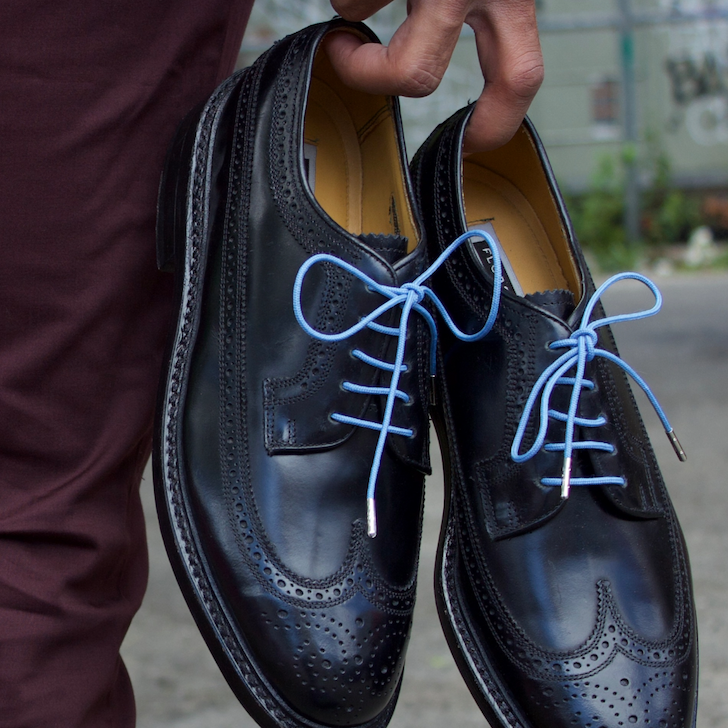 A person holds a pair of black leather brogue shoes with light blue laces, shown close-up outdoors.