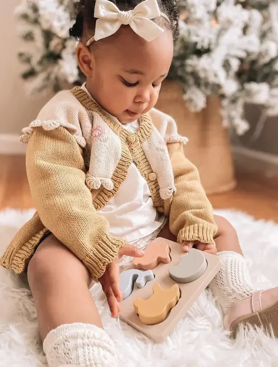 Baby girl wearing a cream cardigan and bow headband sits on a fluffy rug, playing with a wooden shape puzzle.