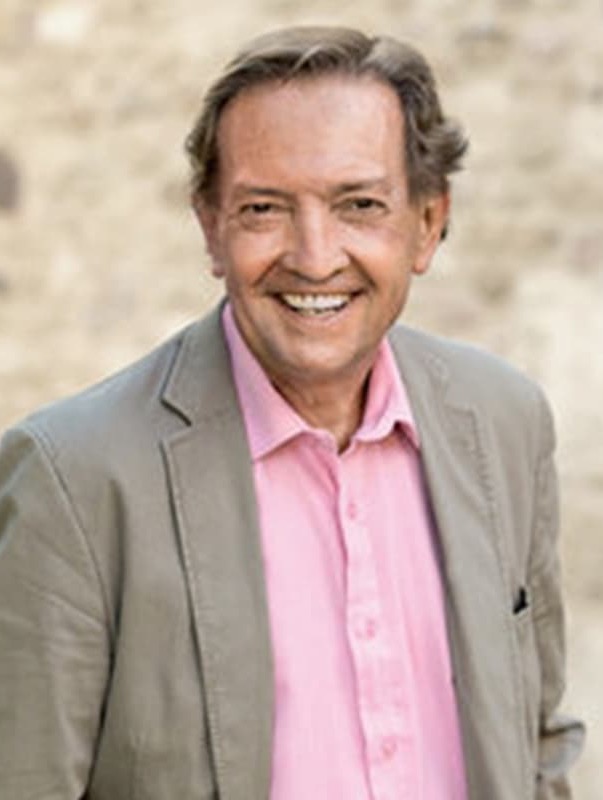 Smiling man wearing a light gray blazer over a pink button-down shirt, posed against a softly blurred stone background. The relaxed portrait conveys warmth and approachability.