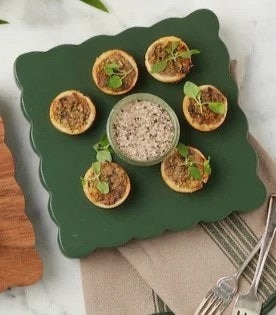 Six bite-sized tartlets topped with fresh herbs surround a bowl of creamy dip on a scalloped dark green serving board, styled on layered neutral linens with silver serving pieces nearby.