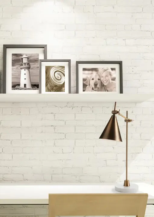 Modern brass desk lamp on a white desk beneath a floating shelf holding framed black-and-white photos against a white brick wall.