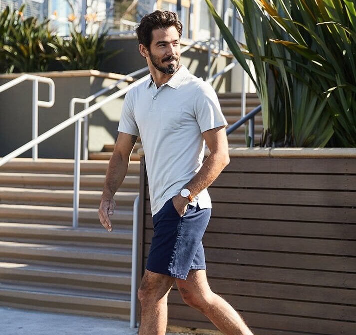 Man walking outdoors on a city sidewalk, wearing a light polo shirt, navy shorts, and sneakers, with stairs, railings, and greenery in the background.
