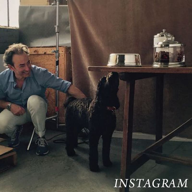 A man crouches next to a black dog beside a table holding a metal dog bowl and two glass containers. The scene appears indoors and has the word "INSTAGRAM" written at the bottom right. Fearrington Village