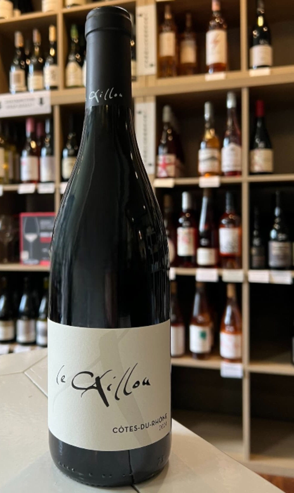 Bottle of Le Grillon Côtes-du-Rhône wine displayed on a counter with shelves of assorted wine bottles in the background.