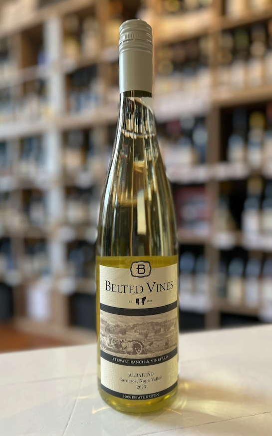 A bottle of Belted Vines Albariño wine stands on a white counter, with shelves of wine bottles softly blurred in the background of a wine shop.