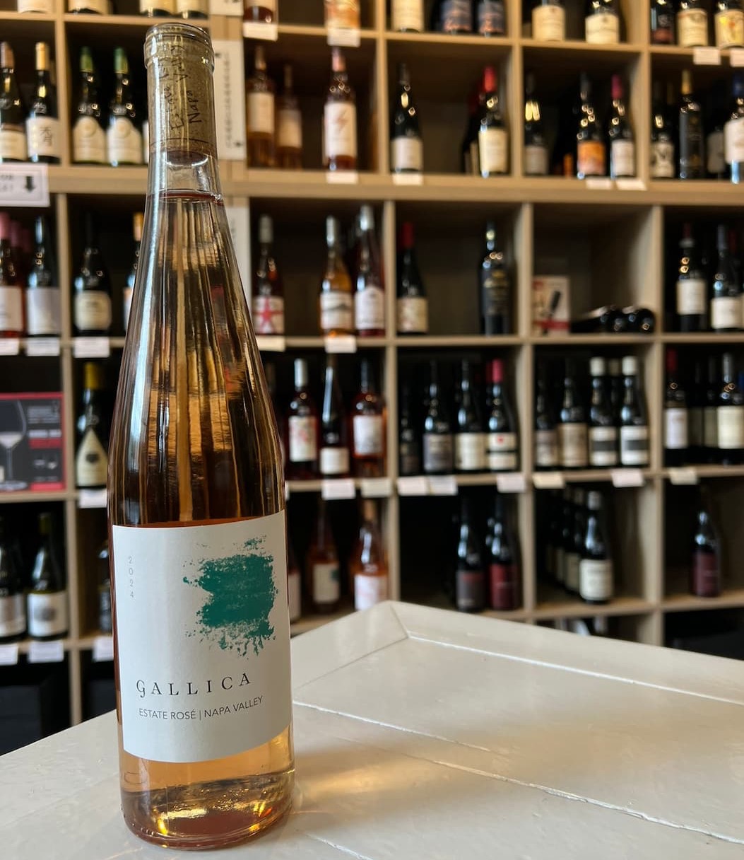 Bottle of Gallica Estate Rosé standing on a white counter in front of a softly blurred wall of wine bottles, highlighting the wine shop setting and the rosé’s warm blush color.