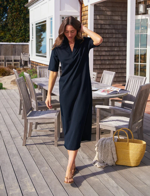 A woman in a relaxed black midi dress walks barefoot across a sunlit wooden deck, brushing her hair back, with outdoor dining furniture, a woven tote, and a coastal-style home in the background.