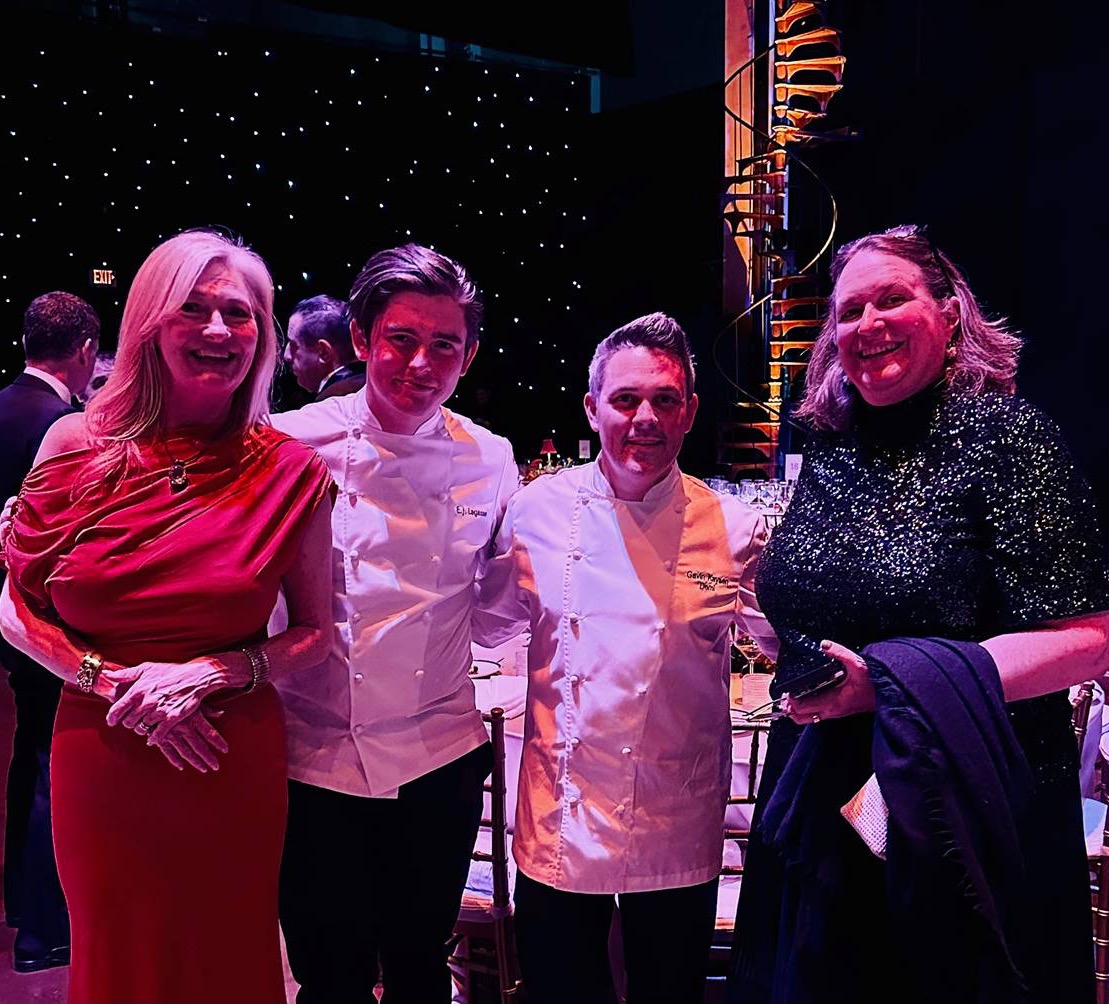 Four people pose together at an elegant event; two women in formal dresses stand on either side of two men wearing chef jackets, with a decorated dining area and star-like lights in the background. Fearrington Village