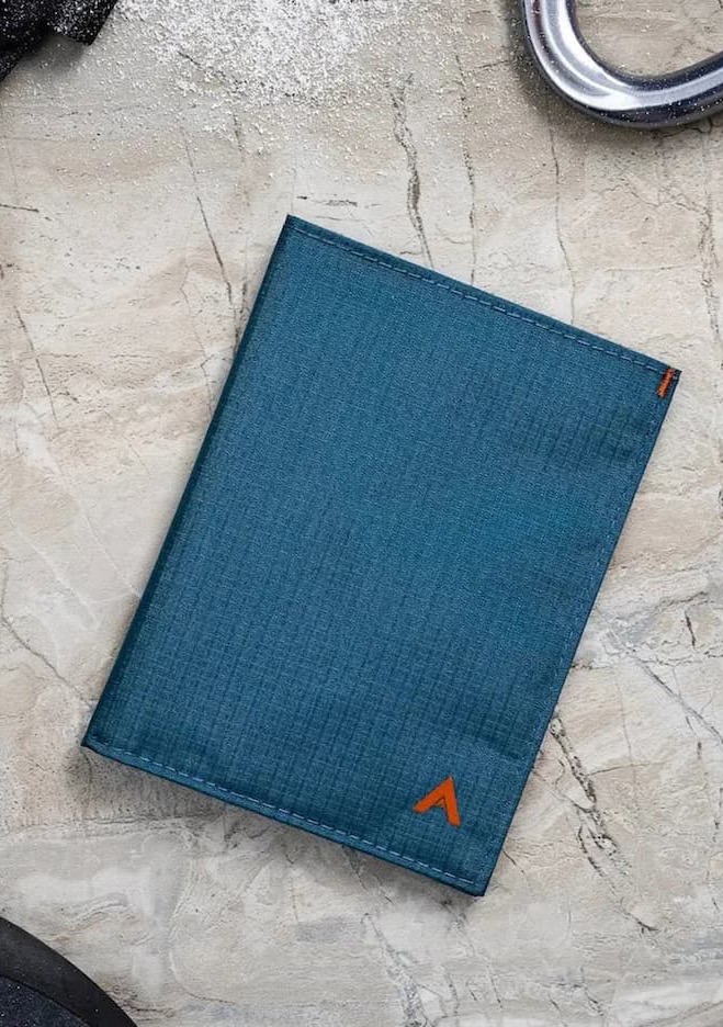 Blue weatherproof passport holder with a minimalist design and orange logo, photographed on a stone surface with outdoor travel gear.