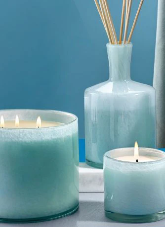A set of pale blue glass candles in two sizes sit beside a matching glass reed diffuser with wooden reeds, arranged on a light surface against a blue background.