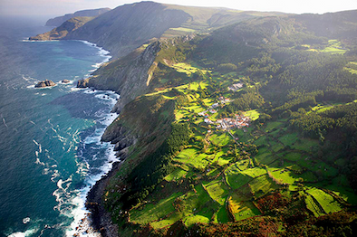 A dramatic coastal landscape where rugged cliffs meet the ocean, with waves crashing along the shoreline and a patchwork of vivid green fields stretching inland. A small village sits nestled among the hills, surrounded by forests and farmland under soft, angled light.