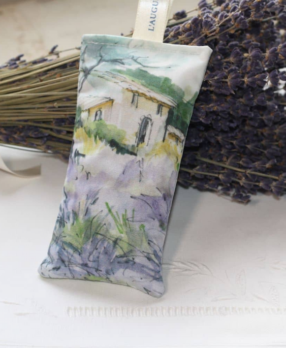 A slender fabric lavender sachet with a loose, sketch-like watercolor of a countryside cottage and wild greenery, propped against a bundle of dried lavender on a light surface.