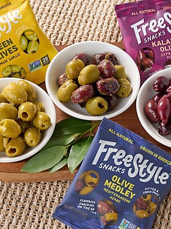Freestyle Snacks olive varieties arranged in small white bowls alongside colorful resealable snack pouches on a woven natural-texture surface. The assortment includes green olives, kalamata olives, and Mediterranean olive medleys, styled as an easy gourmet snack or charcuterie addition.