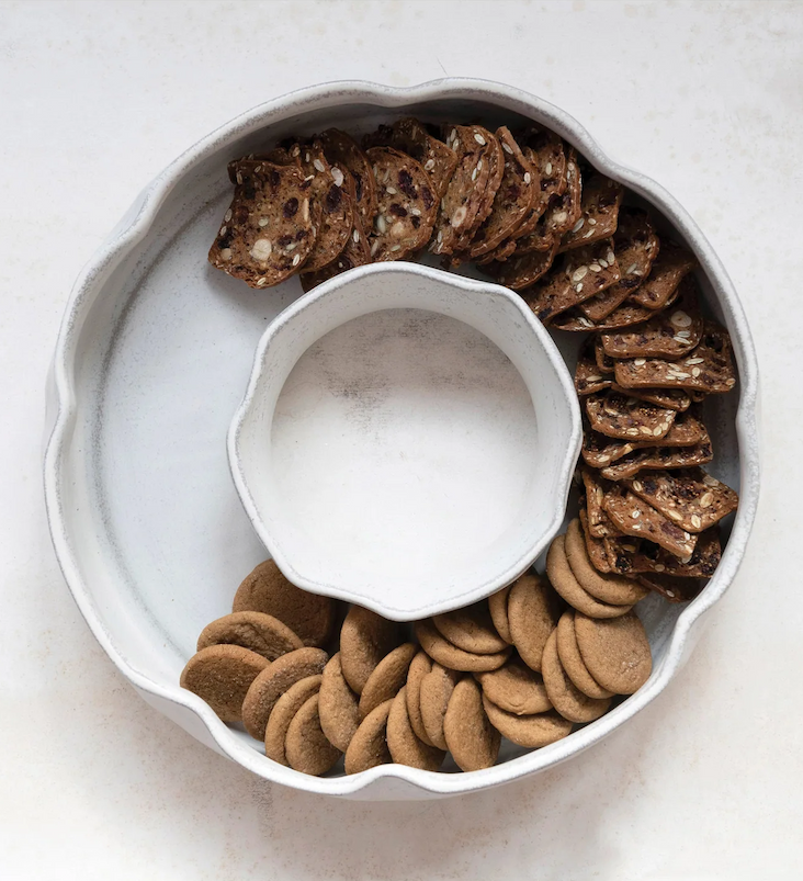 A white stoneware serving dish with a scalloped edge holds an assortment of crackers and cookies arranged in sections around the center.