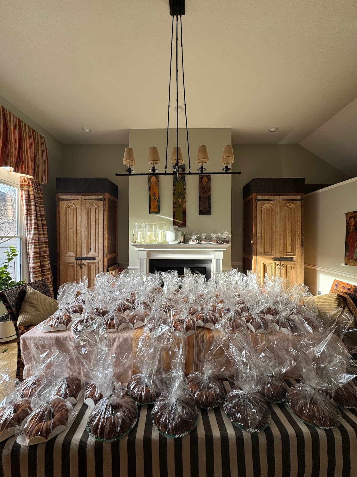 The room is filled with rows of individually wrapped bundt cakes arranged across multiple tables, all gathered under a tall chandelier and framed by warm wood cabinets and soft natural light.