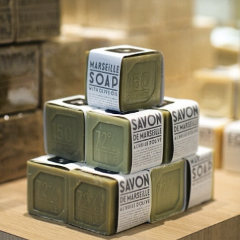 Stacked cubes of Marseille soap wrapped in labeled paper, showcasing olive oil content, arranged on a wooden surface with a softly blurred shop display in the background.