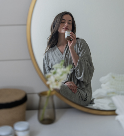 Woman in a soft robe seen through a round mirror, gently holding a small jar near her face as if smelling it, with a calm bedroom setting, muted tones, and a vase of flowers in the foreground.