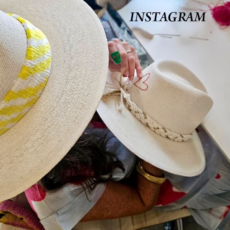 A milliner hand-embroidering an ivory straw hat at a worktable, with another finished hat featuring a yellow patterned band in the foreground, showcasing the craftsmanship behind custom hat making. Fearrington Village.