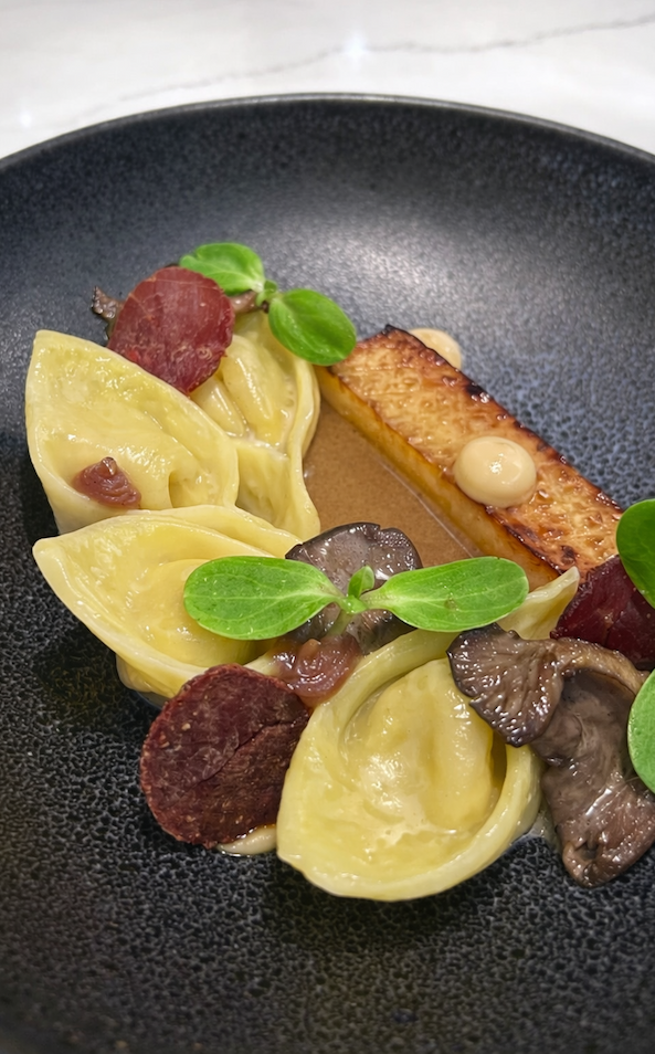 Close-up of a gourmet pasta dish with filled pasta, mushrooms, crisp garnishes, and microgreens arranged on a black ceramic plate.