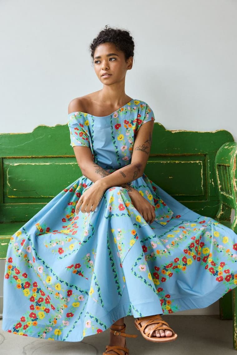 Model seated on a distressed green wooden bench wearing an off-the-shoulder blue dress with colorful floral print, styled with leather sandals against a simple indoor backdrop.