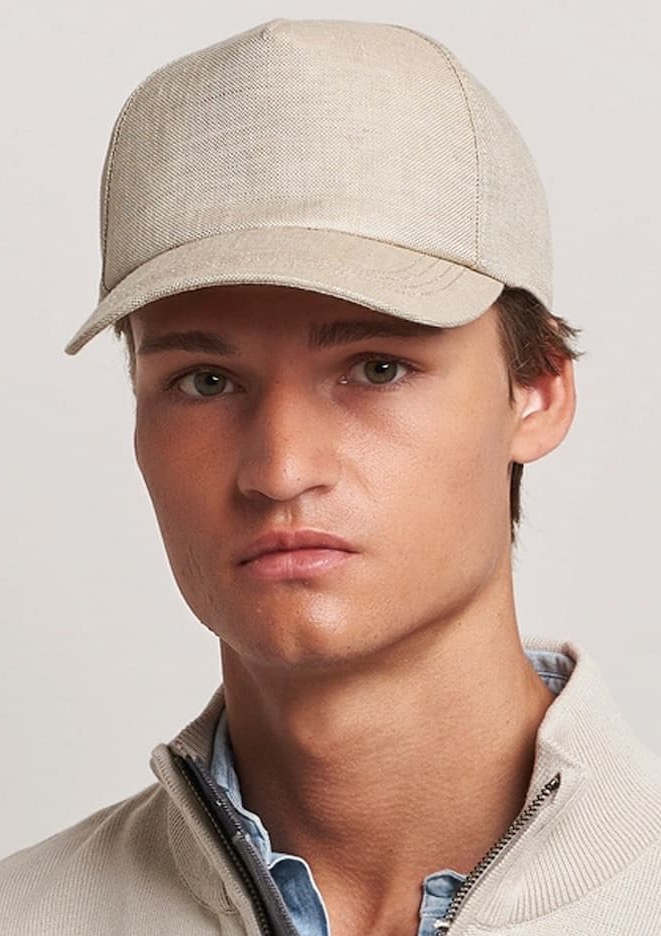 Close-up of a person wearing a beige baseball cap and light jacket, looking straight ahead against a neutral background.