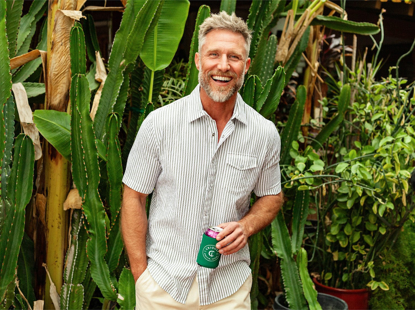 Smiling man in a light striped short-sleeve shirt holding a drink, standing in front of lush greenery and tall cacti.