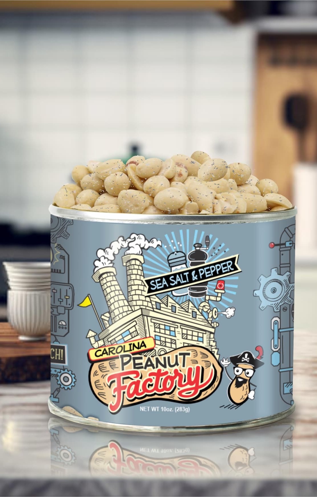 A tin of Carolina Peanut Factory Sea Salt & Pepper peanuts sits on a kitchen countertop, filled with roasted peanuts seasoned with visible flecks of salt and pepper. The playful blue label features illustrated factory machinery, a cartoon peanut mascot, and bold vintage-style lettering. A softly blurred kitchen backdrop adds warmth and highlights the snack as the focal point of the image.