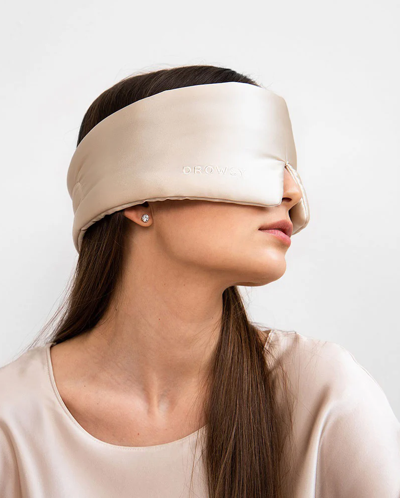 Person in a neutral top wearing a champagne-colored padded sleep mask, shown in profile against a light background.