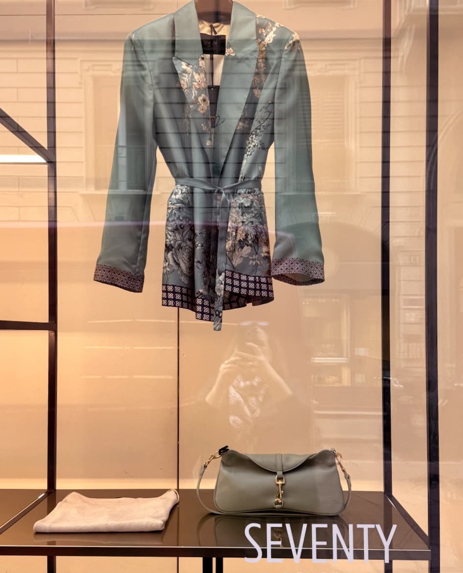 Store window display with a sage-green floral jacket on a hanger above a matching handbag and folded accessory on a black shelf, with street reflections visible in the glass.