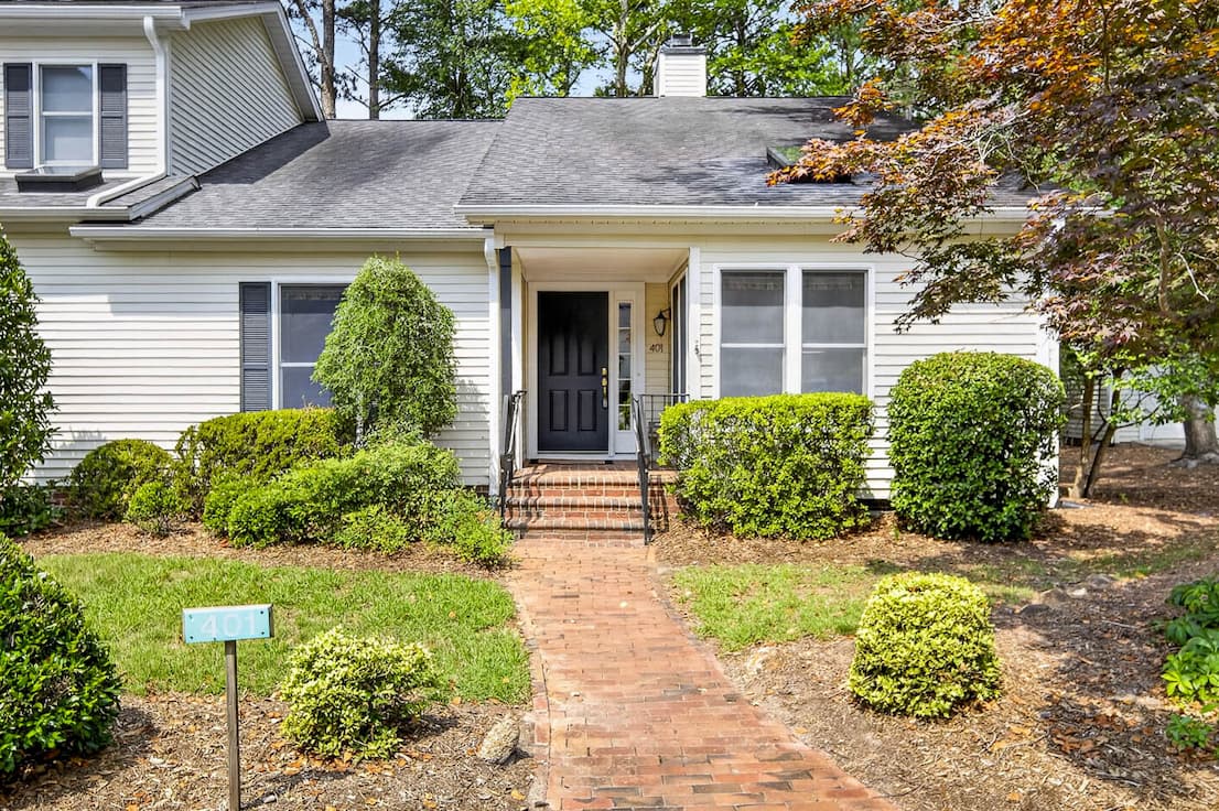 A charming cottage-style home with white siding, a black front door, brick walkway, and beautifully maintained landscaping nestled among mature trees. Fearrington Village.
