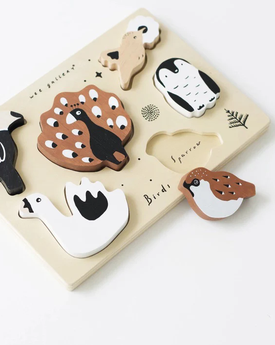 A wooden children’s puzzle features illustrated bird shapes in soft neutral tones, including a swan, penguin, sparrow, and peacock. The pieces sit on a cream-colored board with simple black line drawings and the words “Bird species” printed on the surface.
