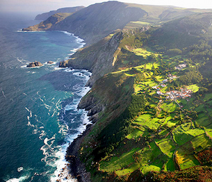 A sweeping aerial view of rugged coastal cliffs meets the ocean, with waves crashing below and sunlit green fields and small villages spread across the rolling hills above.