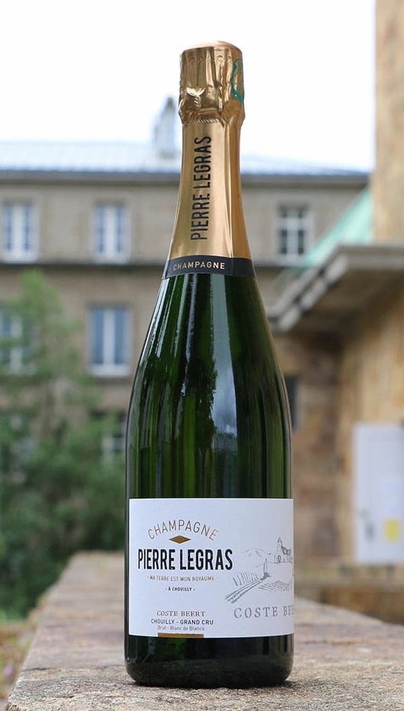 A bottle of Pierre Legras Champagne stands on a stone surface outdoors. The green glass bottle has a gold foil neck and a white label reading “Champagne Pierre Legras, Chouilly Grand Cru,” with a softly blurred building in the background.